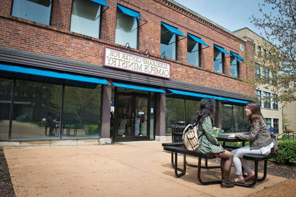 Two people sit at a picnic table outside Eckelkamp Center for Campus Ministry on a sunny day.